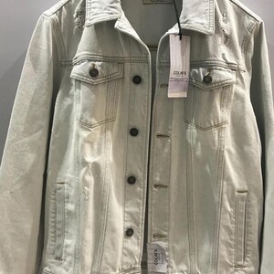 Puede incluir: Una chaqueta vaquera azul claro con detalles desgastados y cierre de botones. La chaqueta tiene dos bolsillos en el pecho y dos bolsillos delanteros. La etiqueta de la chaqueta dice "COLIN'S".