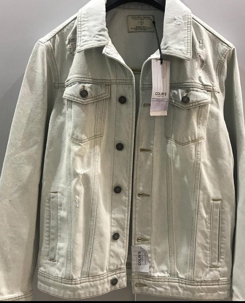 Puede incluir: Una chaqueta vaquera azul claro con detalles desgastados y cierre de botones. La chaqueta tiene dos bolsillos en el pecho y dos bolsillos delanteros. La etiqueta de la chaqueta dice "COLIN'S".