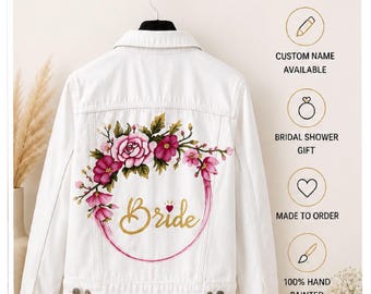 Pode incluir: Casaco de ganga branco com um design floral em torno da palavra "Bride" em letras douradas. O casaco apresenta uma grinalda floral rosa e bordô. O texto na imagem inclui "Custom Name Available", "Bridal Shower Gift" e "Made to Order".