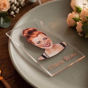 Könnte beinhalten: Eine transparente Platzkarte aus Acryl mit einer Aquarellillustration einer rothaarigen Frau und dem Namen "Clara Rose" in goldfarbenen Buchstaben. Die Platzkarte liegt auf einem grünen Teller, mit Blumen und einer Gabel im Hintergrund.