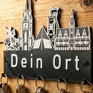 Könnte beinhalten: Ein schwarzer Schlüsselhalter mit weißem Skyline-Design und dem Text "Dein Ort". Der Schlüsselhalter hat mehrere Haken für Schlüssel und ist auf einer Holzoberfläche montiert.