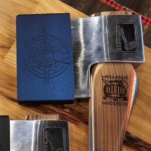 May include: A collection of axes with wooden handles and metal blades. The axes have various designs etched into the metal and the handle. One axe has the text "WELLMAN AXE & SAW WORKS WOODSON" on the handle. The axes are displayed on a wooden surface.
