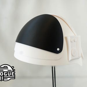 May include: A white astronaut helmet featuring a smooth matte finish, a curved black visor, and a rectangular side panel with circular details. The logo ROGUE is printed in the bottom left corner in a black and white graphic design.