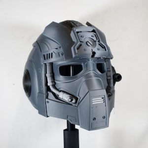 Puede incluir: Un casco táctico de color gris oscuro mate que presenta una visera frontal prominente, un protector nasal con rejillas de ventilación horizontales y un riel para accesorios en la parte superior. Incluye componentes circulares laterales y un tubo flexible.