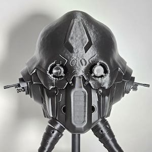 May include: A dark gray, futuristic helmet with intricate mechanical details. The helmet features circular eye-like components, a central breathing apparatus, and side-mounted gun-like structures. The design suggests a science fiction or cosplay theme.