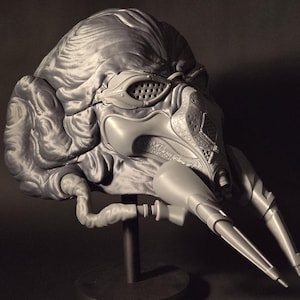 Könnte beinhalten: Eine graue, detaillierte Skulptur eines futuristischen Helms mit komplizierten Designs und einer strukturierten Oberfläche. Der Helm hat ein Visier mit kleinen Löchern und zwei lange, spitze Anhänge.
