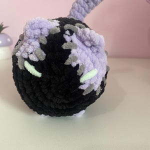May include: A handmade crochet cat toy in black, lavender, and gray. The cat toy has a round body with a tail and small ears. The toy is made of soft, textured yarn and has small green accents.