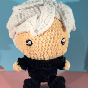 Puede incluir: Una muñeca de peluche hecha a mano con una cara de color canela claro, ojos negros y cabello blanco. La muñeca lleva un atuendo negro. La muñeca está sentada sobre una superficie rosa con un fondo azul.