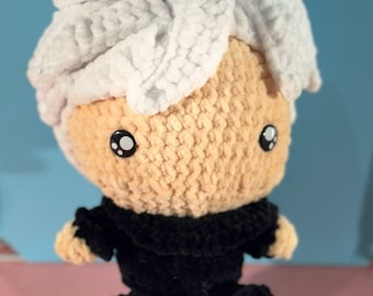 May include: A handmade plush doll with a light tan face, black eyes, and white hair. The doll is wearing a black outfit. The doll is sitting on a pink surface with a blue background.
