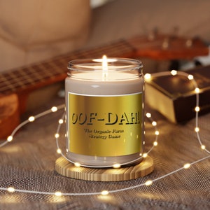 Peut inclure: Une bougie allumée dans un pot en verre transparent avec une étiquette dorée qui dit "OOF-DAH! The Organic Farm Strategy Game." La bougie repose sur un petit dessous de verre en bois, entourée de guirlandes lumineuses. Une guitare et un livre sont en arrière-plan.