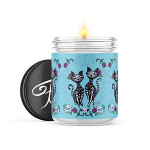 May include: A blue glass candle jar with a white lid. The jar has a design of two black cats with sugar skulls on their faces. The cats are surrounded by flowers and vines. The candle is lit.