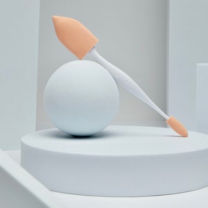 Pode incluir: Um pincel de maquiagem branco com uma ponta de esponja de cor pêssego claro. O pincel está apoiado em uma esfera branca e uma plataforma branca. O nome da marca "SAYEBI" está impresso no cabo.