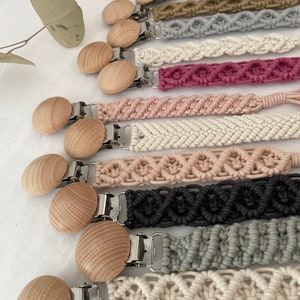 Peut inclure: Un ensemble de dix attaches-sucette en bois avec des lanières crochetées de différentes couleurs. Les attaches sont en bois naturel et ont un fermoir en métal argenté. Les lanières sont en fil de coton et présentent différents motifs, notamment des chevrons, des cœurs et des cercles.