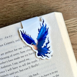 May include: A bookmark featuring a blue bird design with outstretched wings, placed on the pages of a book. The bird has a gradient of colors, transitioning from light peach to deep blue. The bookmark is white with the bird design.