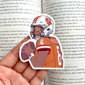 May include: A sticker featuring an illustration of a football player in an orange and white uniform, holding a football. The player wears a helmet with a logo and the number 6 on the jersey. The sticker is on a book page.