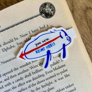 May include: A white and blue bookmark with the text "You were RIGHT HERE!" and a red arrow. The bookmark is shaped like a stylized pig and is placed on an open book.