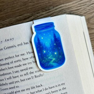 May include: A blue jar-shaped bookmark featuring a forest scene with trees, a winding path, and glowing lights. The bookmark is placed on the pages of a book, with text visible. The bookmark is made of a glossy material.