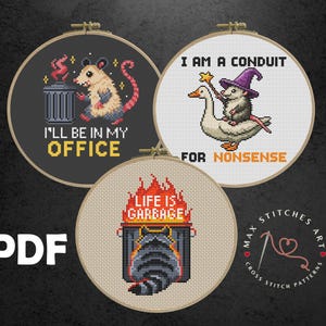 Puede incluir: Tres patrones de punto de cruz en aros de bordado. Uno presenta una zarigüeya junto a un cubo de basura con el texto "I'LL BE IN MY OFFICE". Otro muestra una zarigüeya montada en un ganso con el texto "I AM A CONDUIT FOR NONSENSE". El tercero tiene un mapache y el texto "LIFE IS GARBAGE."