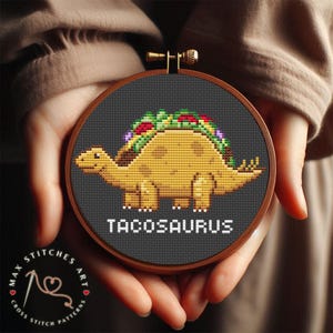 Puede incluir: Un patrón de punto de cruz con un dinosaurio de dibujos animados que lleva una cáscara de taco como caparazón. El dinosaurio es marrón y amarillo, y la cáscara de taco está llena de ingredientes coloridos. El texto "TACOSAURUS" está escrito debajo del dinosaurio.
