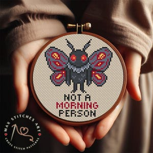 May include: A cross-stitch embroidery in a wooden hoop features a moth design with red and gray wings and the text "NOT A MORNING PERSON." The artwork is a cross-stitch pattern.