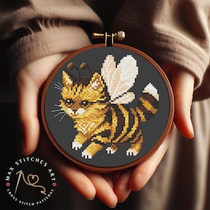 May include: Cross-stitch embroidery of a cat with bee wings and stripes, set in a wooden hoop. The cat is yellow and black, with white wings and a white-tipped tail, set against a dark gray background. The text "MAX STITCHES ART" is visible.