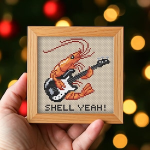 May include: A square cross-stitch embroidery in a light wood frame featuring an orange shrimp playing a black and white electric guitar. The design is stitched on a cream-colored fabric background with the black text SHELL YEAH! at the bottom.
