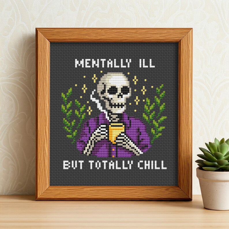 May include: Cross-stitch artwork in a wooden frame. The design shows a skeleton in a purple shirt, holding a yellow cup, and smoking. Text reads "MENTALLY ILL BUT TOTALLY CHILL" in white block letters. Green leaves and yellow stars surround the skeleton.