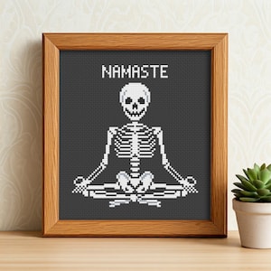 Op de afbeelding: Een ingelijst kruissteekontwerp met een skelet in een yogapose met het woord "NAMASTE" erboven.
