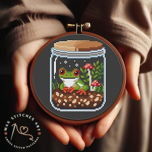 Op de afbeelding: Een kruissteekpatroon van een groene boomkikker die in een glazen pot zit met paddenstoelen en groen. De pot is omgeven door een bruine houten ring.