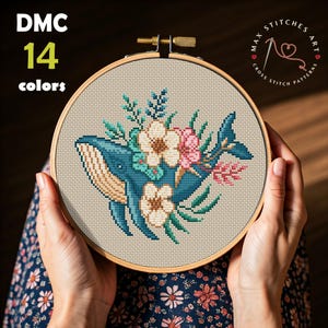 Puede incluir: Un patrón de punto de cruz de una ballena azul con flores rosas, blancas y verdes. El patrón está en un aro de bordado de madera y está siendo sostenido por las manos de una persona. El texto "MAX STITCHES ART CROSS STITCH PATTERNS" está en el aro.