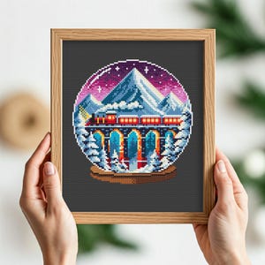 Puede incluir: Un patrón de punto de cruz de un globo de nieve con un tren que viaja a través de una cordillera. El globo de nieve es azul y blanco con un cielo morado y rosa. El tren es rojo y amarillo.