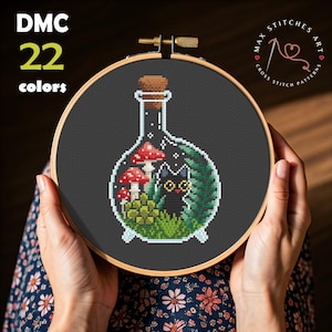 May include: Cross-stitch pattern featuring a black cat inside a bottle with red mushrooms and green foliage. The design is held in a wooden embroidery hoop. Text on the image reads "DMC 22 colors" and "MAX STITCHES ART CROSS STITCH PATTERNS".