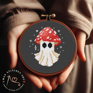 Può includere: Uno schema di punto croce con un fungo rosso e bianco con un viso di fantasma. Il fungo è su uno sfondo nero con stelle bianche.
