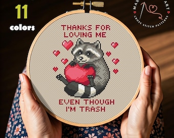 Może przedstawiać: Tamborek do haftu krzyżykowego z szopem trzymającym serce, otoczonym sercami. Tekst brzmi "THANKS FOR LOVING ME EVEN THOUGH I'M TRASH." Projekt wykorzystuje 11 kolorów DMC. Tamborek jest trzymany przez ręce.