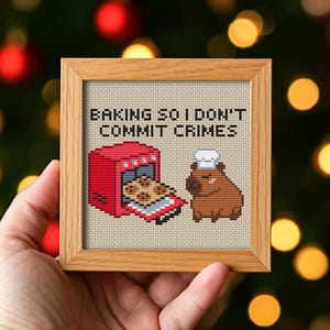 May include: A framed cross-stitch with the text "BAKING SO I DON'T COMMIT CRIMES." The image shows a red oven with cookies and a brown capybara wearing a chef's hat. The frame is made of light brown wood.