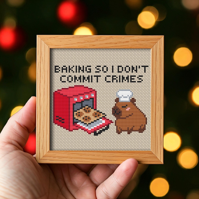 May include: A framed cross-stitch piece with the text "BAKING SO I DON'T COMMIT CRIMES." The image features a red oven with cookies and a brown capybara wearing a chef's hat. The frame is made of light brown wood.