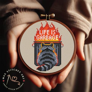 Könnte beinhalten: Ein Kreuzstich-Stickrahmen mit einem Design eines brennenden Mülleimers und dem Text "LIFE IS GARBAGE". Das Design verwendet Grau-, Schwarz-, Orange- und Rottöne. Der Rahmen wird in den Händen einer Person gehalten.