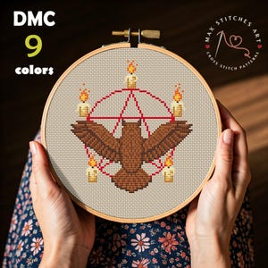 May include: A cross-stitch embroidery hoop featuring a brown bird design with a red pentagram and candles. The text "DMC 9 colors" is visible. The hoop is held by hands, and the design is set against a beige fabric.