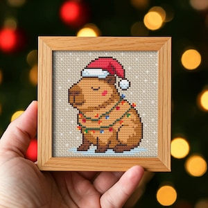 Può includere: Un punto croce incorniciato con un capibara marrone che indossa un cappello da Babbo Natale e luci natalizie. L'opera d'arte è ambientata su uno sfondo beige chiaro con un sottile effetto neve, incorniciata in una cornice quadrata in legno naturale.