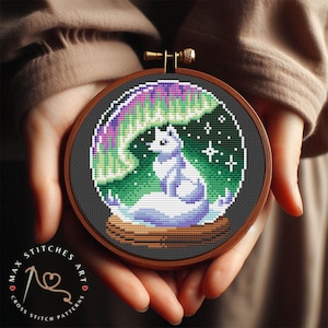May include: A white fox sitting under a colorful aurora borealis in a dark green forest. The fox is surrounded by stars and is sitting on a wooden log. The image is in a round embroidery hoop.