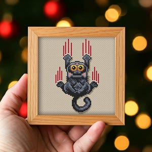 May include: A square cross-stitch embroidery in a light wood frame featuring a dark gray creature with large yellow eyes and a long tail. The creature is depicted with four sets of vertical red claw marks above its paws on a beige fabric background.