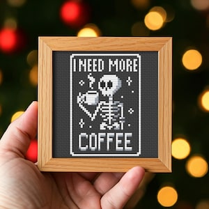 Puede incluir: Un diseño de punto de cruz enmarcado con un esqueleto sosteniendo una taza de café con el texto "Necesito más café".