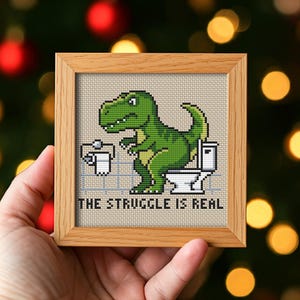 May include: Cross-stitch artwork in a wooden frame. The image shows a green dinosaur on a toilet with the text "THE STRUGGLE IS REAL". The background is a light beige with a grid pattern, and the frame is a light brown colour.