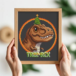 May include: A cross-stitch pattern of a green Christmas tree wearing a brown dinosaur hat. The dinosaur is in a circle with an orange outline and the text "TREE REX" is below the circle.