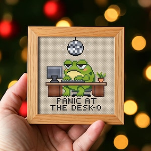 Op de afbeelding: Kruissteekkunstwerk in een houten lijst. De afbeelding toont een groene kikker aan een bureau met een computer, een discobal en de tekst "PANIC AT THE DESK-O". De achtergrond is neutraal.