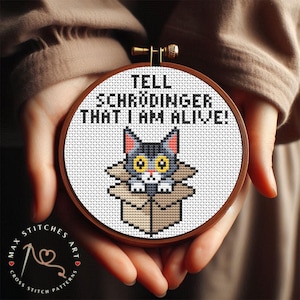 May include: A cross-stitch embroidery hoop with a brown wooden frame. The design features a grey cat in a cardboard box with the text "TELL SCHRÖDINGER THAT I AM ALIVE!". The bottom left corner has the text "MAX STITCHES ART".