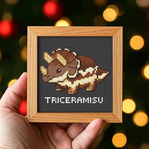 Könnte beinhalten: Ein gerahmtes Kreuzstichbild mit einem braunen und beigen Triceratops-Design auf einem dunkelgrauen Hintergrund. Der Holzrahmen umgibt das Kunstwerk, darunter das Wort "TRICERAMISU". Das Kunstwerk wird vor einem verschwommenen Hintergrund gehalten.