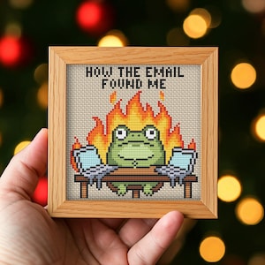 Könnte beinhalten: Ein quadratisches Kreuzstich-Stickbild in einem hellen Holzrahmen zeigt einen grünen Frosch, der an einem Schreibtisch mit zwei Laptops sitzt. Hinter dem Frosch sind orange und gelbe Flammen zu sehen. Der Text darüber lautet "HOW THE EMAIL FOUND ME".