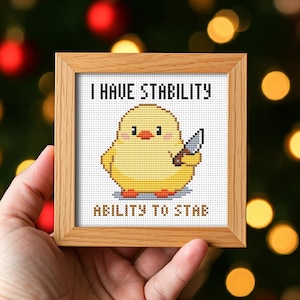 May include: A framed cross-stitch depicting a yellow chick with a knife. The text says "I HAVE STABILITY" and "ABILITY TO STAB". The frame is made of light brown wood.