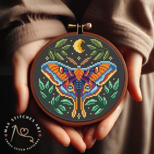 Könnte beinhalten: Ein Kreuzstichmuster mit einem bunten Falter mit einer Sichelmond und Blättern auf einem dunklen Hintergrund. Das Muster befindet sich in einem hölzernen Stickrahmen.